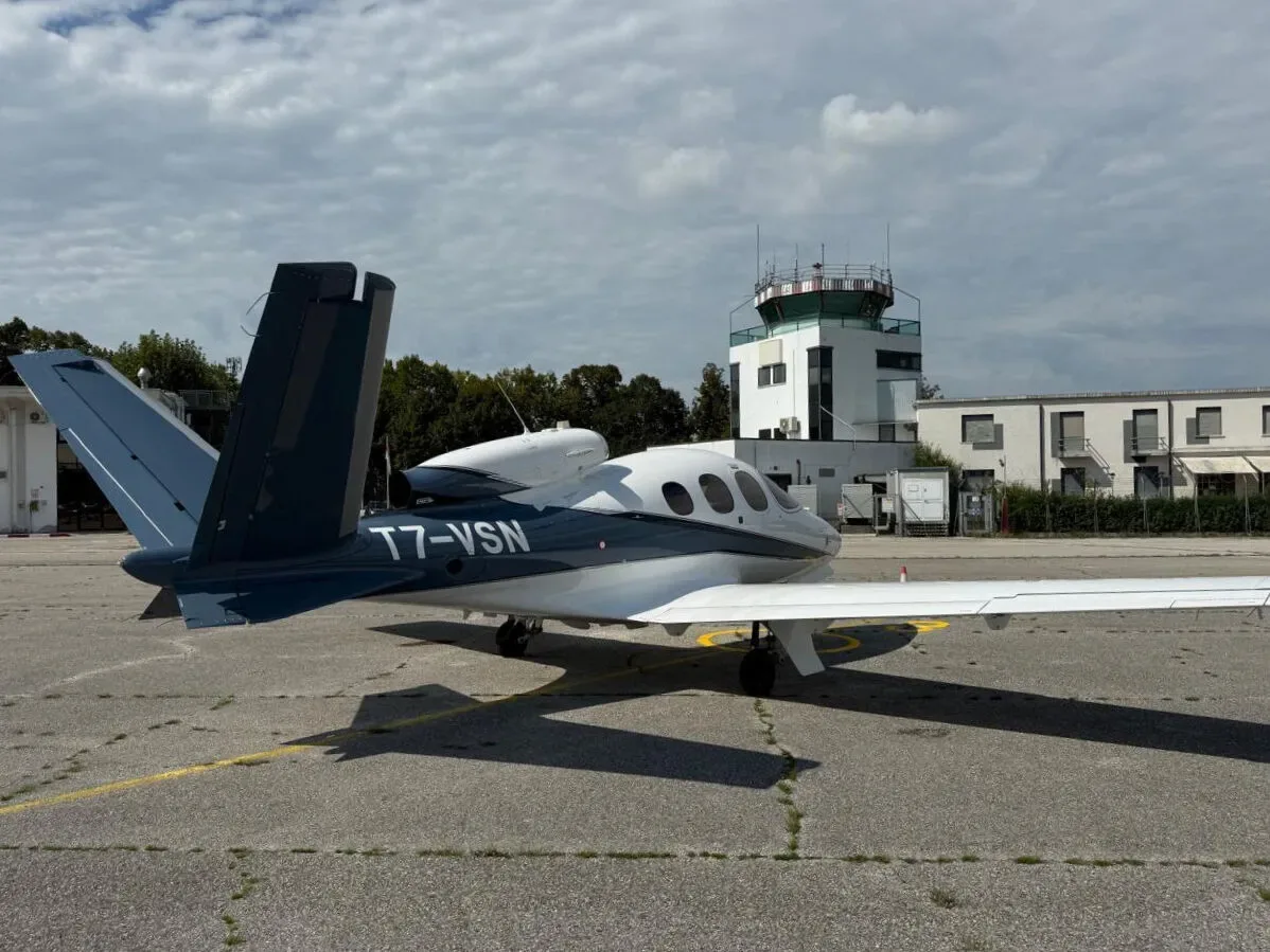 A white and dark blue Cirrus Vision Jet (T7-VSN) parked on an airfield with an air traffic control tower in the background.
