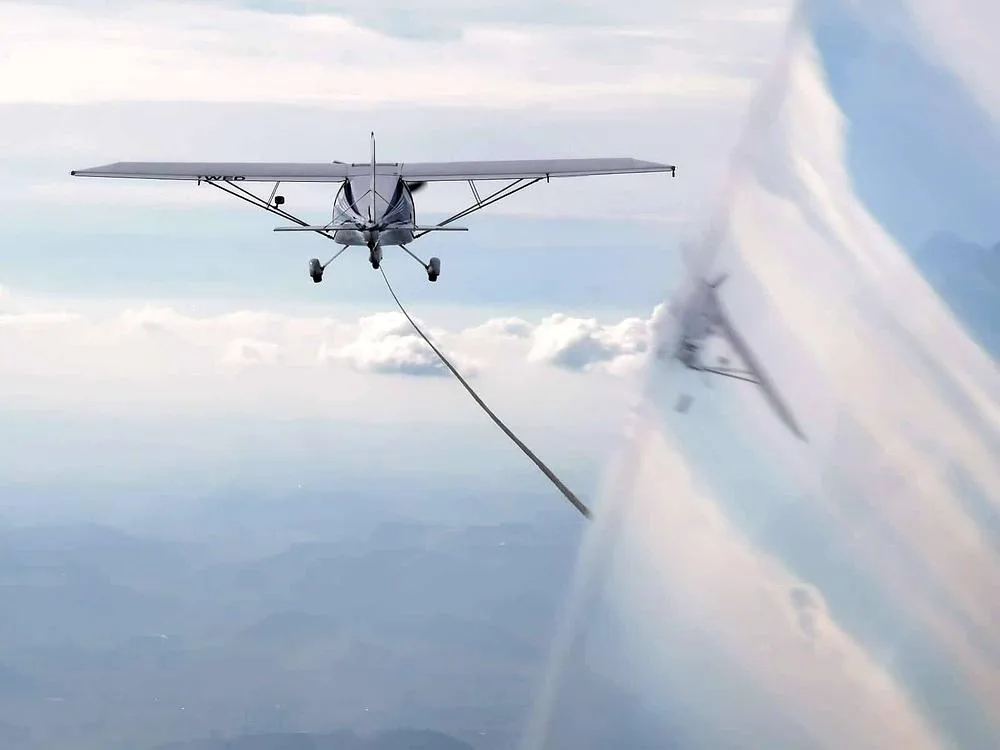 A grey tow plane is seen from the perspective of a glider it is towing, against a cloudy sky with distant land.