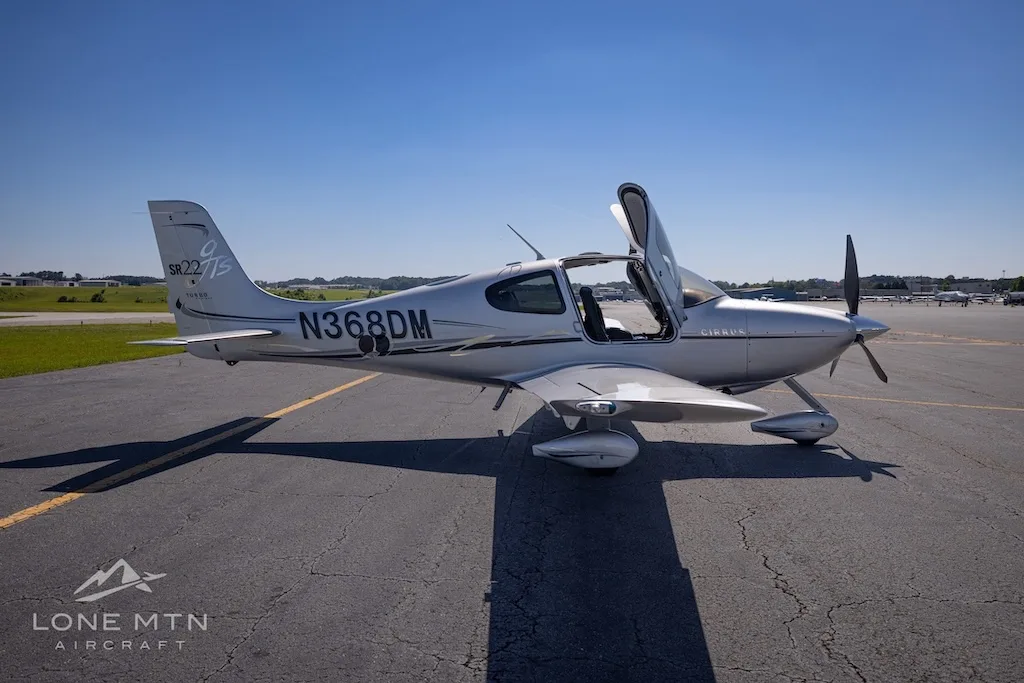 A silver Cirrus SR22 Turbo aircraft (N368DM) with its doors open, parked on an asphalt runway under a clear blue sky.