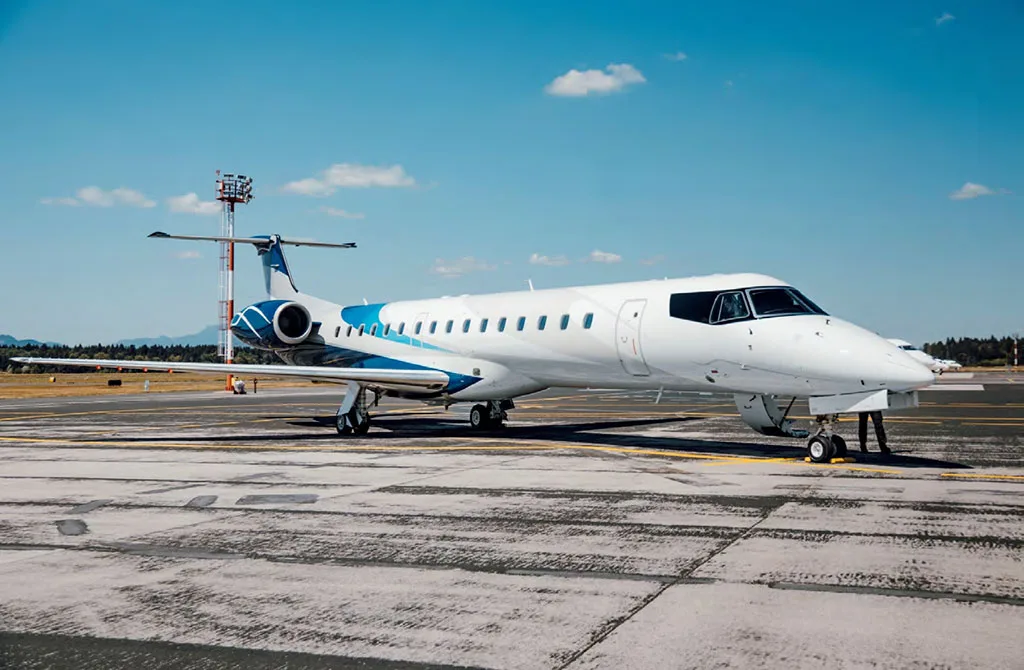A white and blue Embraer ERJ 145 regional jet parked on an airport tarmac under a clear blue sky.
