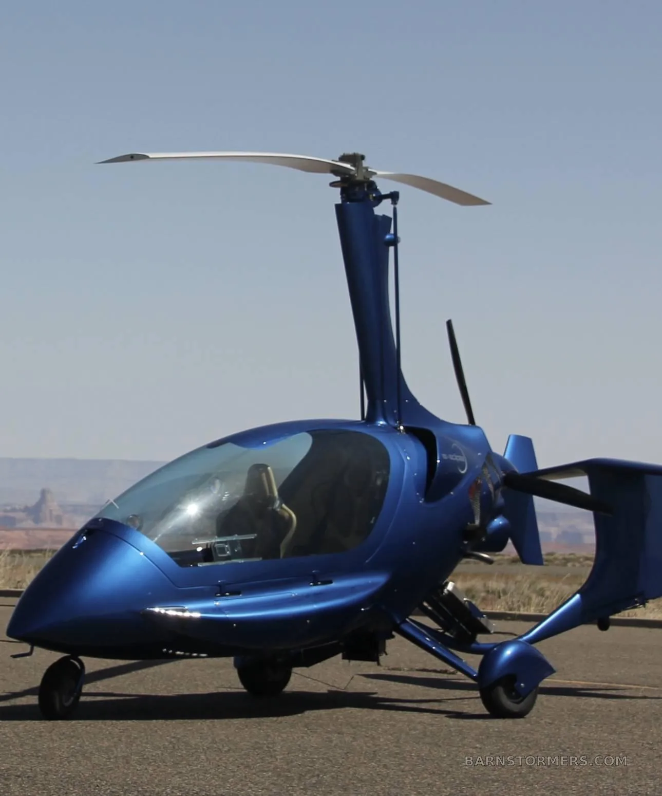 A blue enclosed gyrocopter with a single main rotor and tail propeller sits on an asphalt runway with desert mountains in the background.