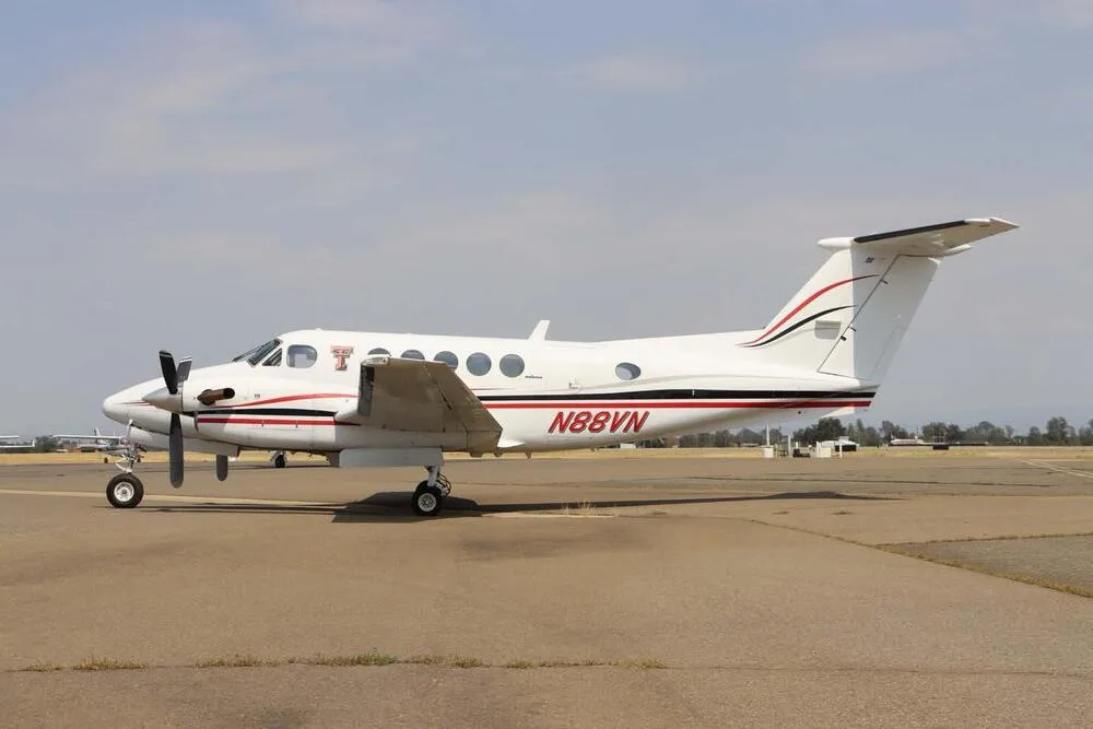 A white Beechcraft King Air N88VN with red and black stripes on the tarmac under a partly cloudy sky.