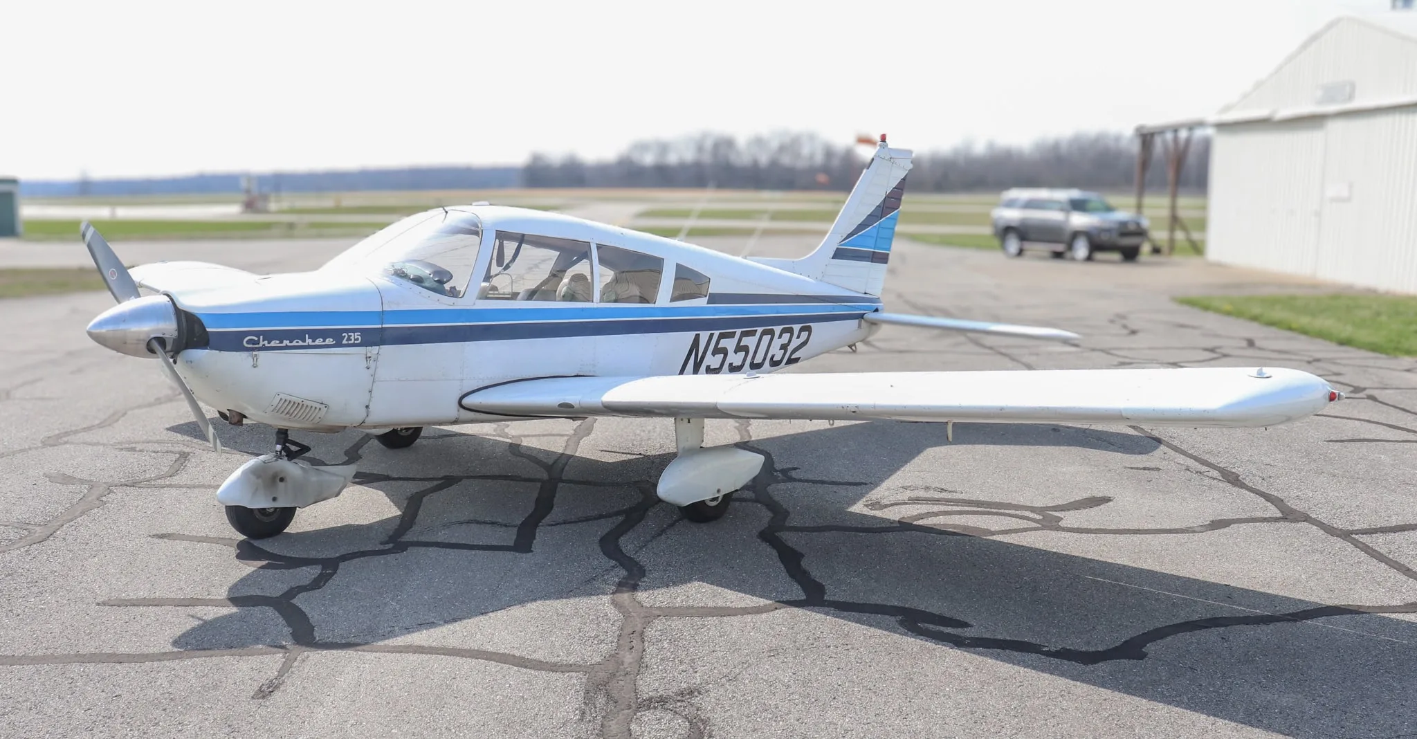 A side view of the white Piper Cherokee 235 aircraft with blue stripes and registration N55032, parked on an asphalt ramp.