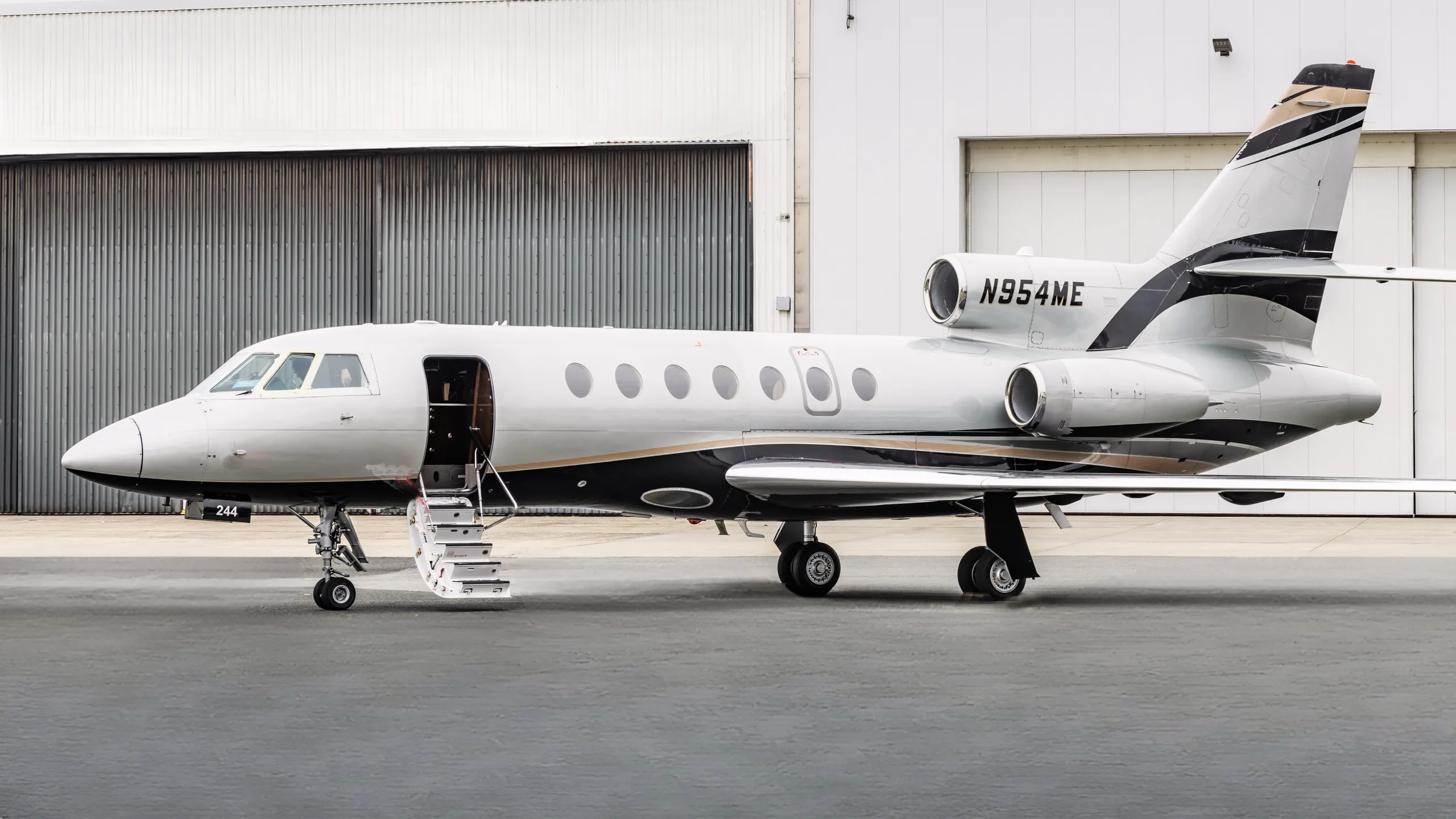 A silver and black Dassault Falcon business jet with gold accent stripes, parked on an apron in front of a hangar with its airstairs deployed.