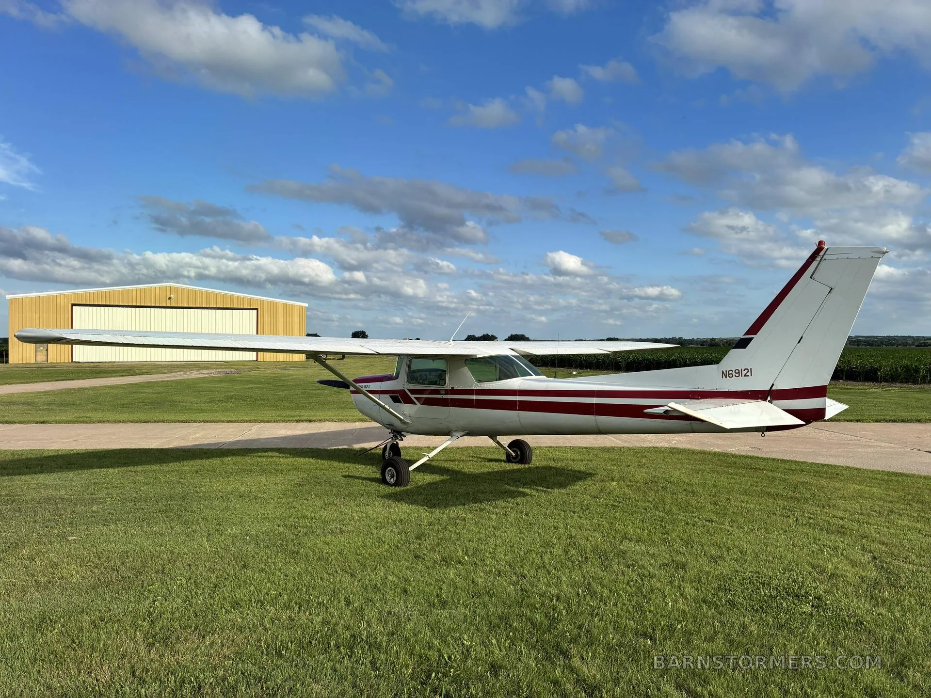 A white and red Cessna 172 aircraft is parked on a grassy field with a yellow hangar in the background.