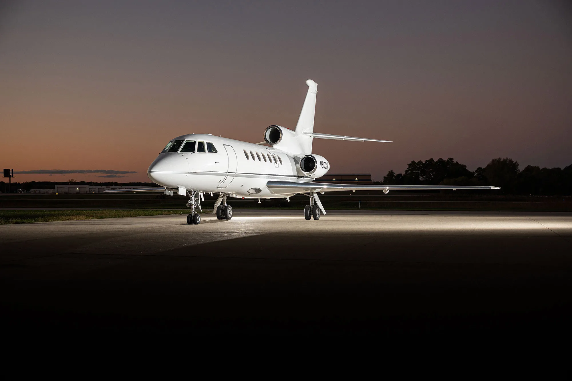 A white Dassault Falcon 900EX business jet (N803NL) parked on an airfield at sunset, front-quarter view.