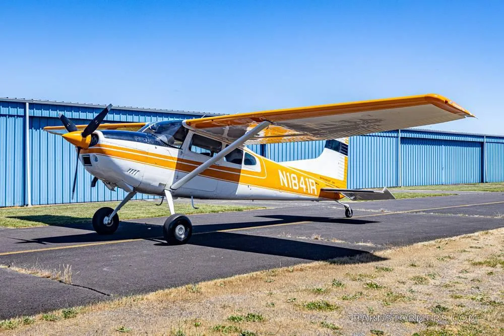 A white and orange single-engine propeller aircraft with tail number N1841R parked on an asphalt ramp in front of blue hangars.