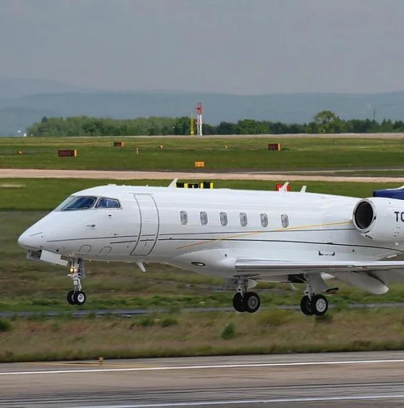 A white business jet with blue tail accents is seen on a runway, with its landing gear extended.