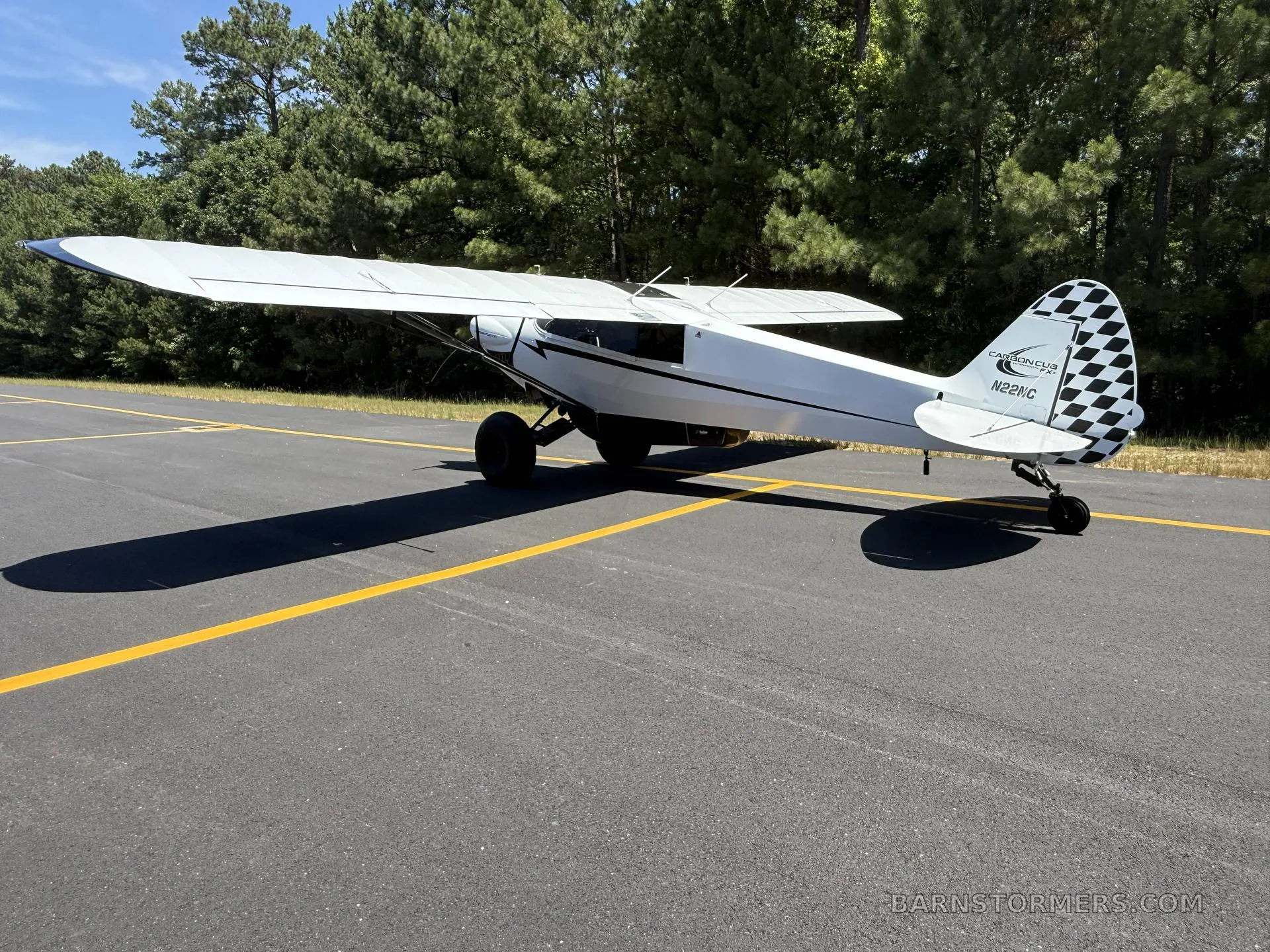 A white Carbon Cub FX-3 aircraft with black accents and a checkered tail is parked on an asphalt runway with trees in the background.