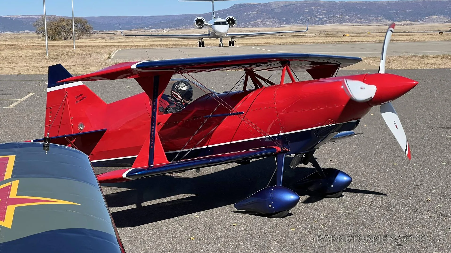 A red and blue biplane with a pilot in the cockpit is parked on an airfield, with a white business jet in the background.