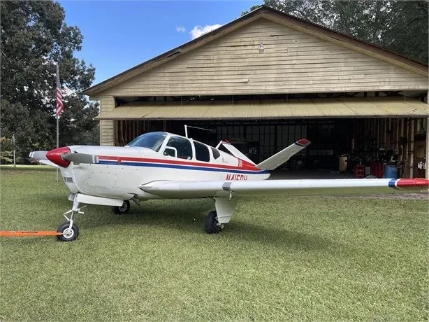 A white and red Beechcraft Bonanza N415DH parked on grass in front of a wooden hangar.