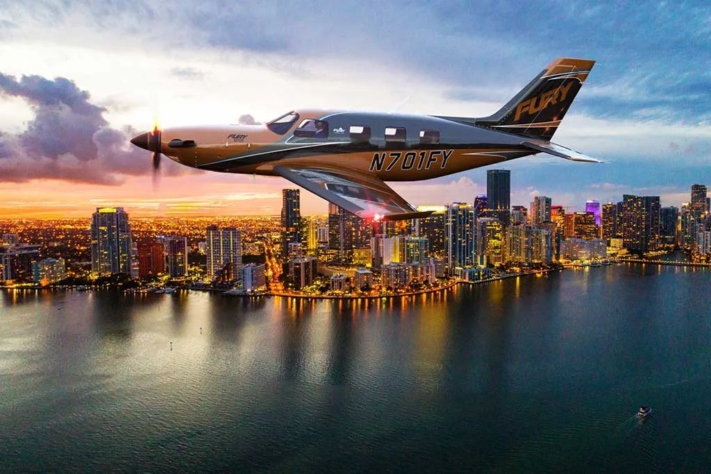 A bronze and black single-engine turboprop aircraft (N701FY) with 'FURY' on the tail, flying over a city skyline at sunset.