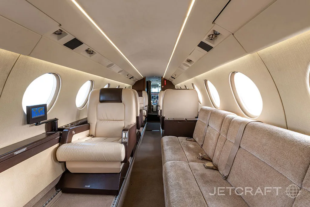 A long view of the business jet cabin interior with beige leather seats, a sofa, and large oval windows.
