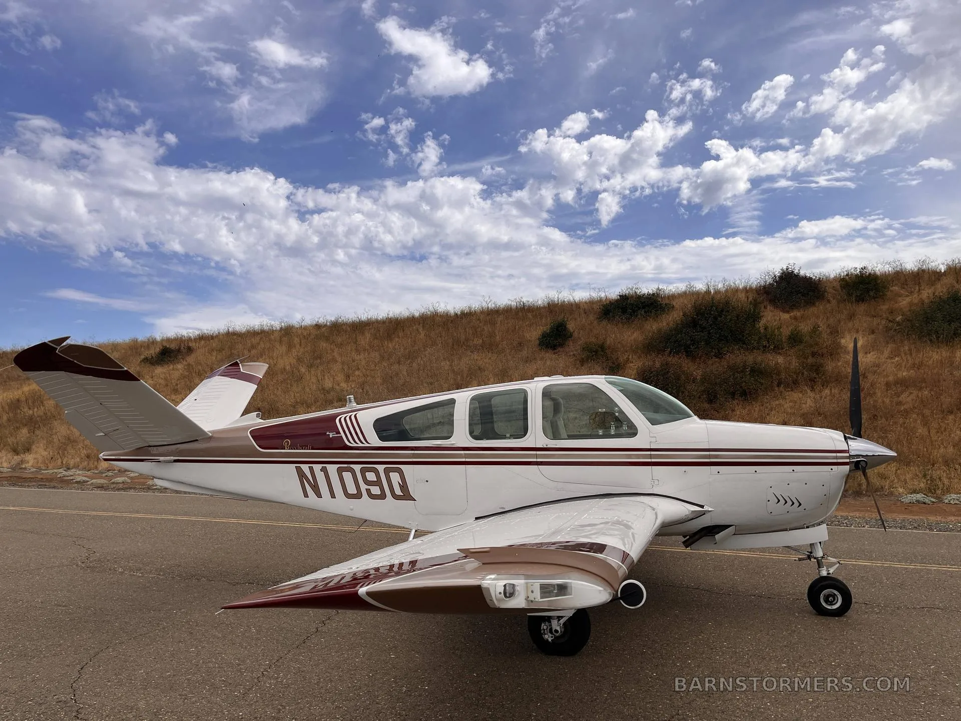 A white and brown Beechcraft Bonanza with registration N109Q, parked on an asphalt surface with a dry grassy hill under a blue sky.
