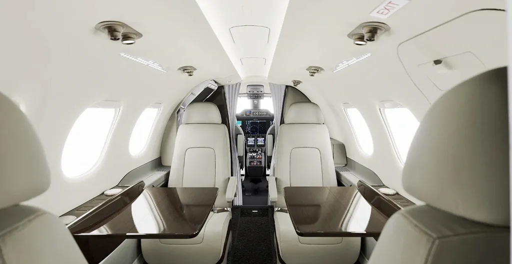 The interior cabin of a private jet with light-colored seats and tables, looking towards the cockpit.