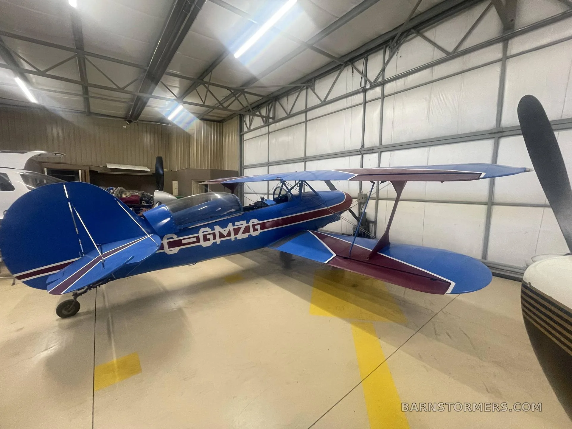 A rear-quarter view of a blue and red biplane with registration C-GMZG parked in a hangar next to another aircraft's propeller.