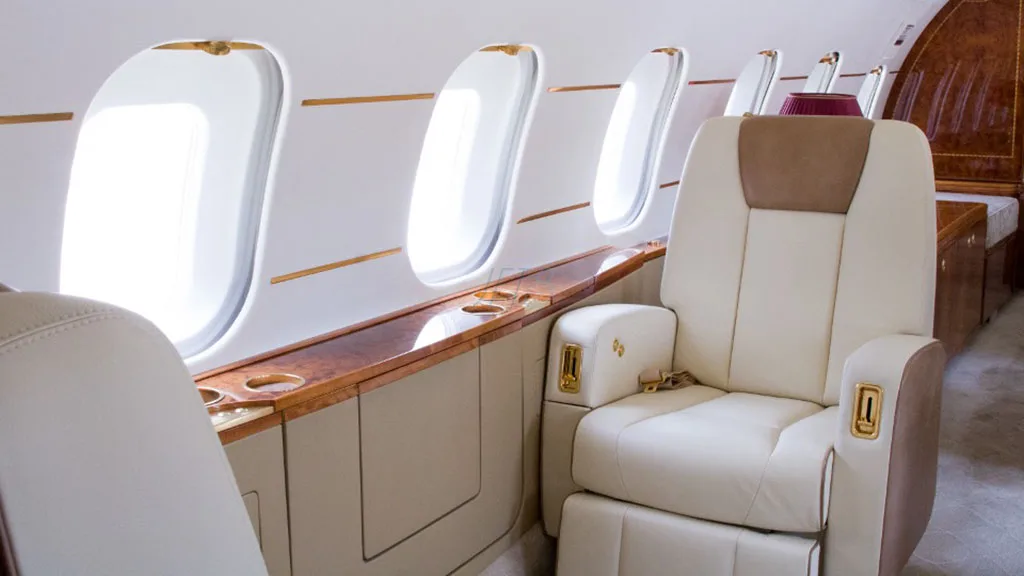 A close-up of a cream-colored leather seat with a brown headrest next to large windows in an aircraft cabin.