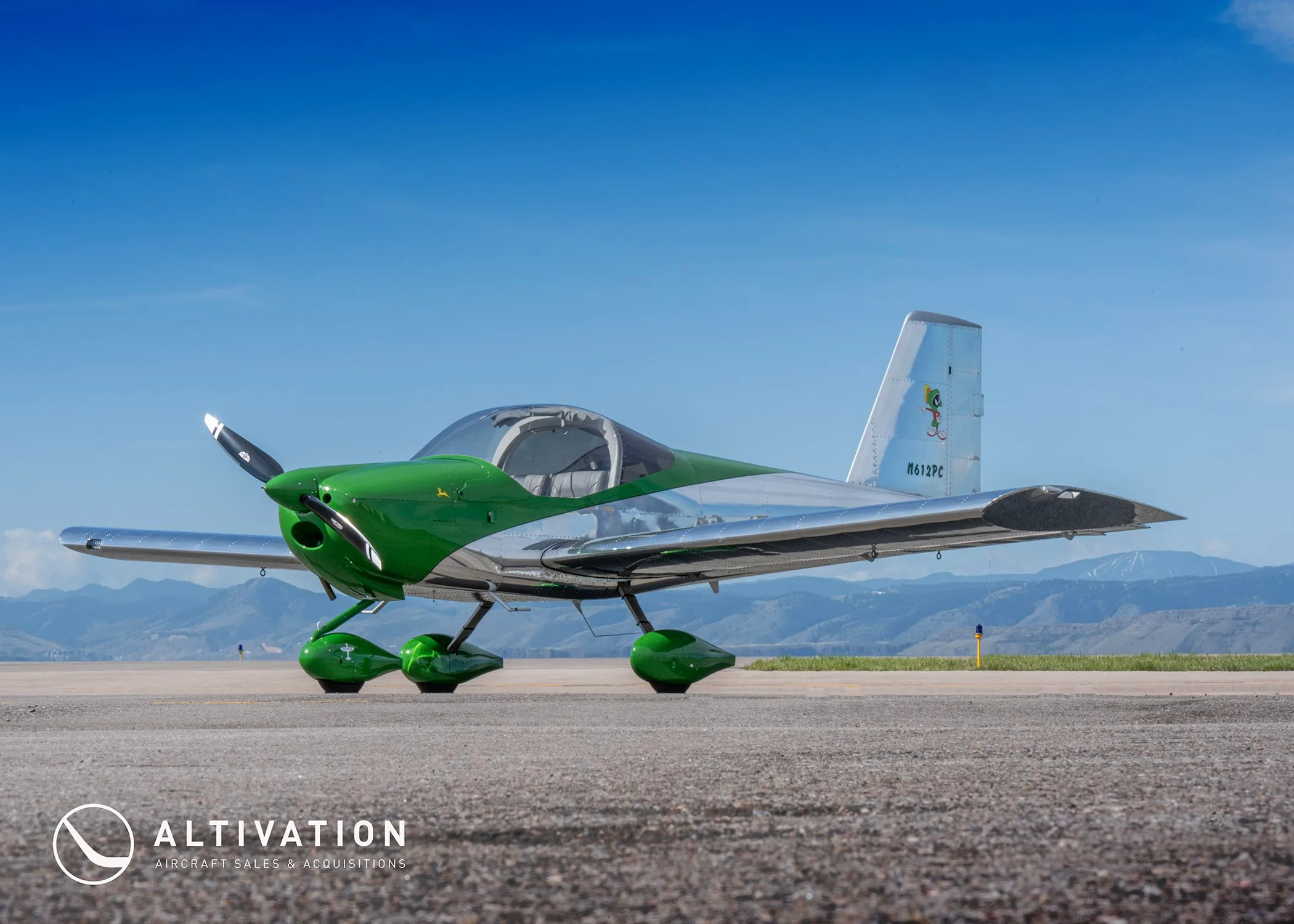 A green and polished aluminum Vans RV-12 aircraft, registration N612PC, parked on an airfield with mountains in the background.