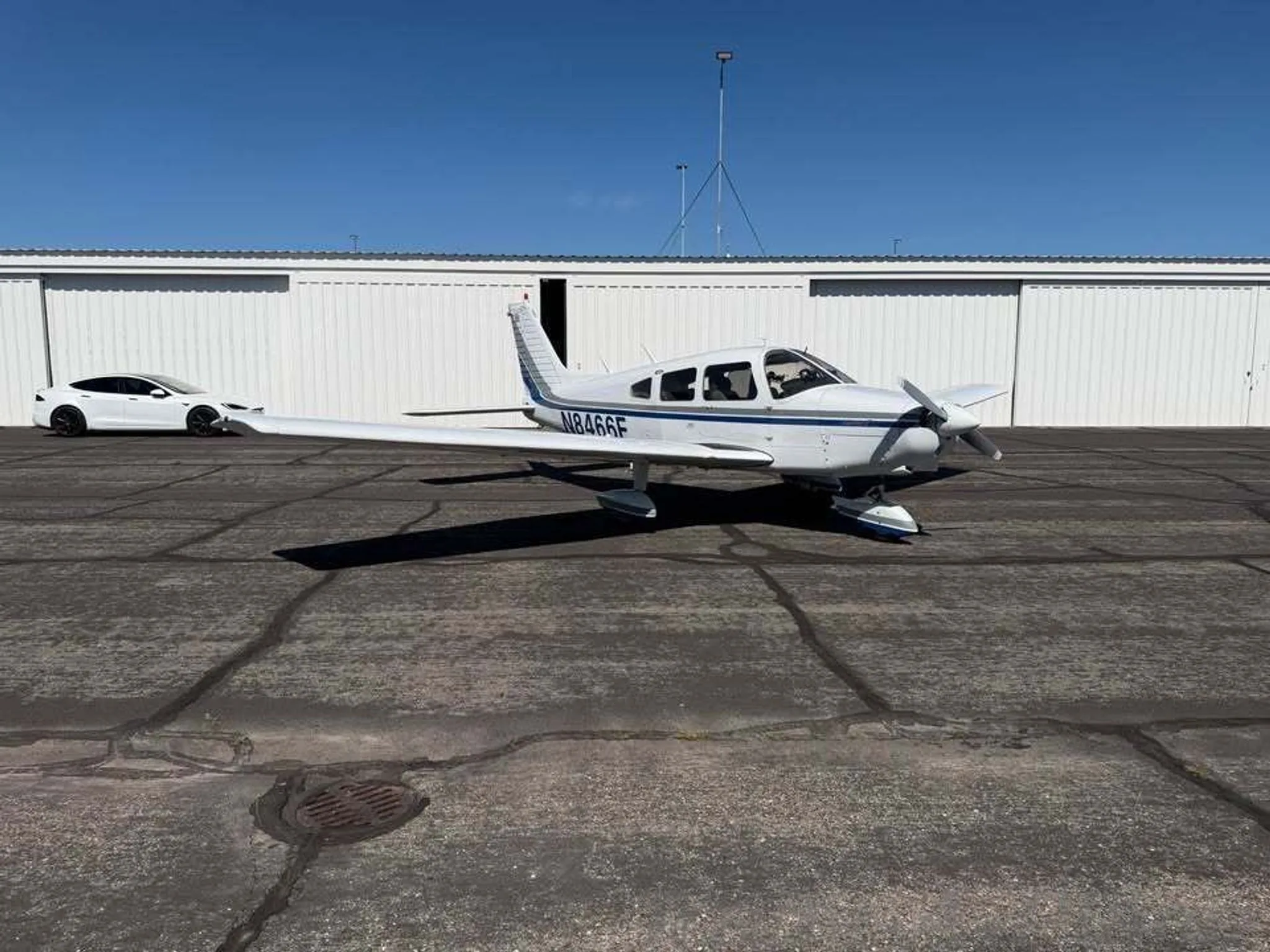 A white Piper Warrior II aircraft with blue and gray stripes and registration N8466F parked on an asphalt apron with a white car in the background.