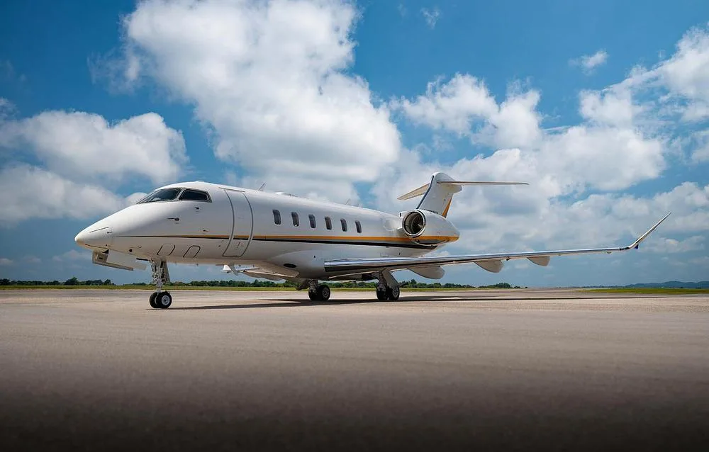 A white business jet with yellow and grey stripes is parked on an airfield under a partly cloudy sky.