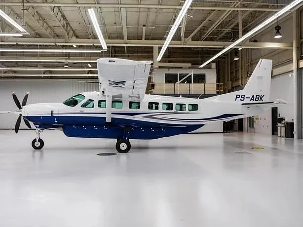 A white and blue Cessna Caravan aircraft with registration PS-ABK is parked inside a brightly lit hangar.
