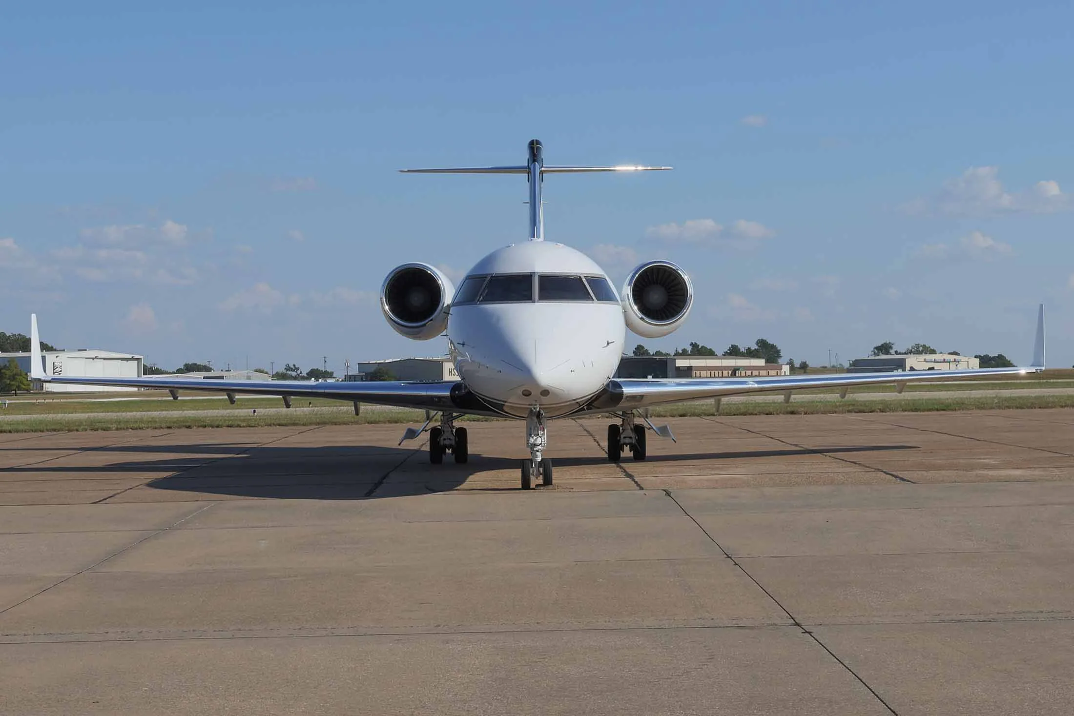 A white Bombardier Challenger business jet seen head-on, parked on an airport tarmac with blue sky and scattered clouds.