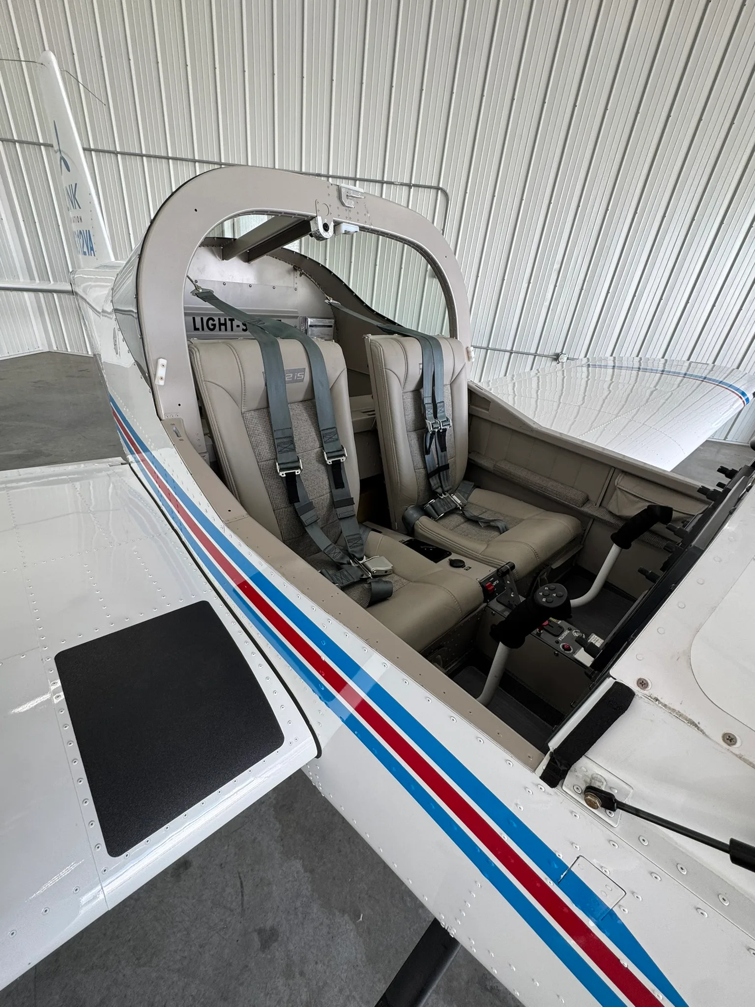The two-seat interior of a light-sport aircraft, showing beige seats with grey fabric inserts and seatbelts.