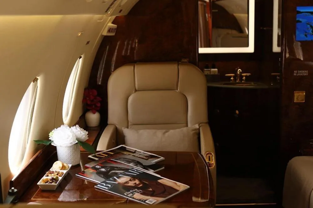 A close-up of a private jet's interior showing a beige seat, a polished table with magazines, and a lavatory entrance.