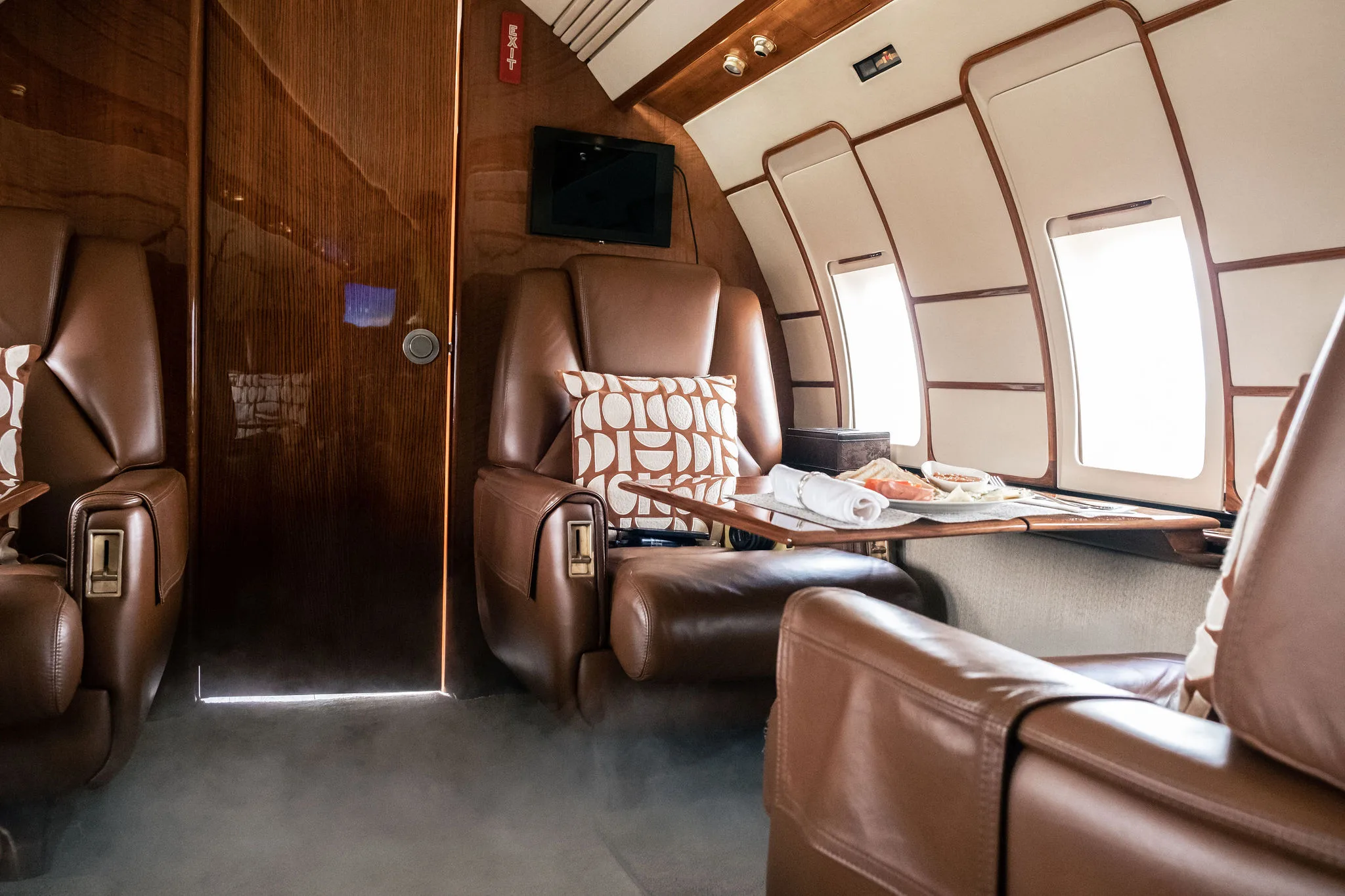 Private jet cabin with brown leather seats, a table set with snacks, and a wooden partition.