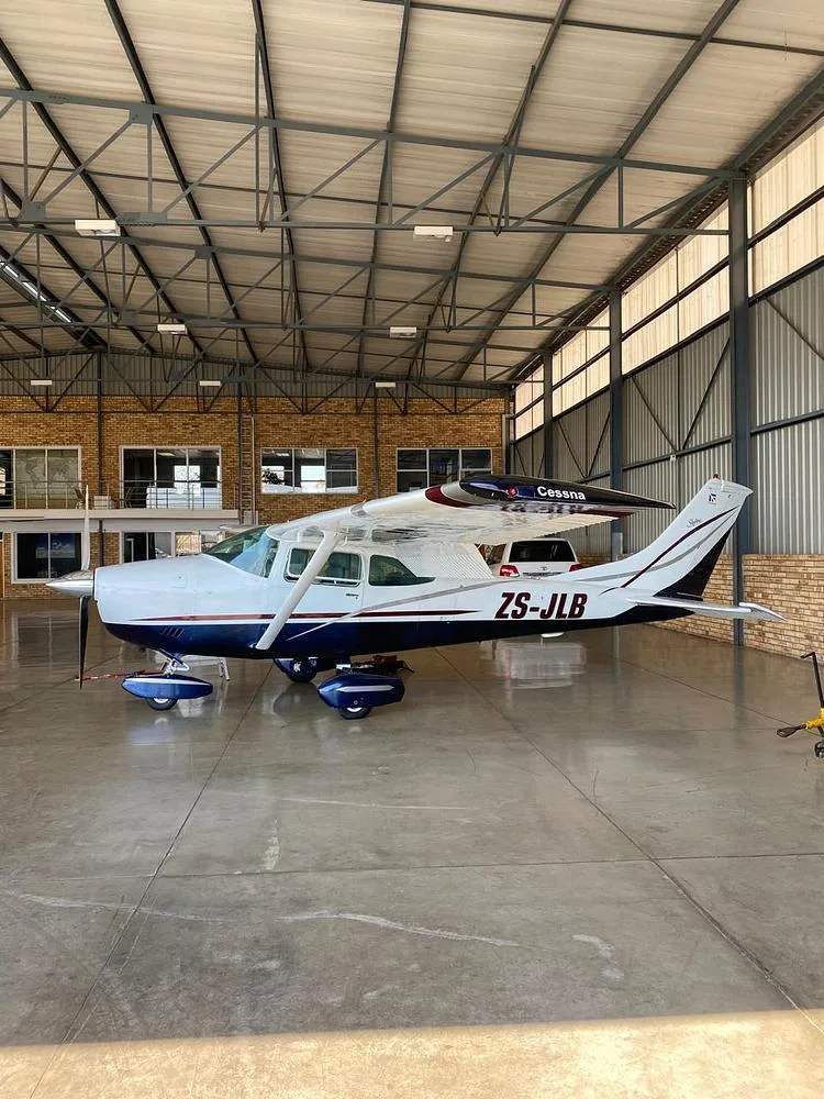 A white and blue Cessna ZS-JLB aircraft is parked inside a large hangar with a high metal roof structure.
