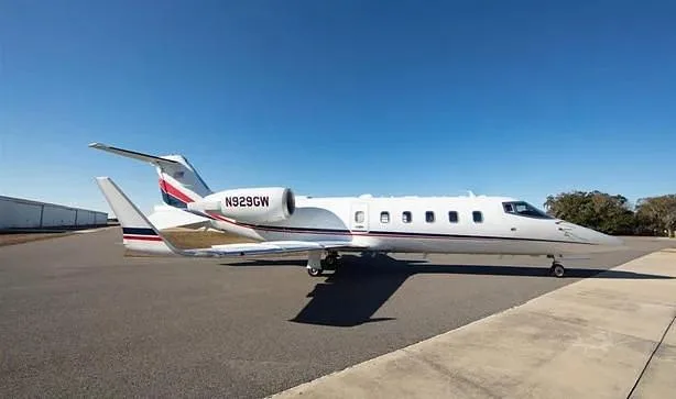 A white Learjet with red and blue stripes, registration N929GW, parked on an asphalt tarmac under a clear blue sky.