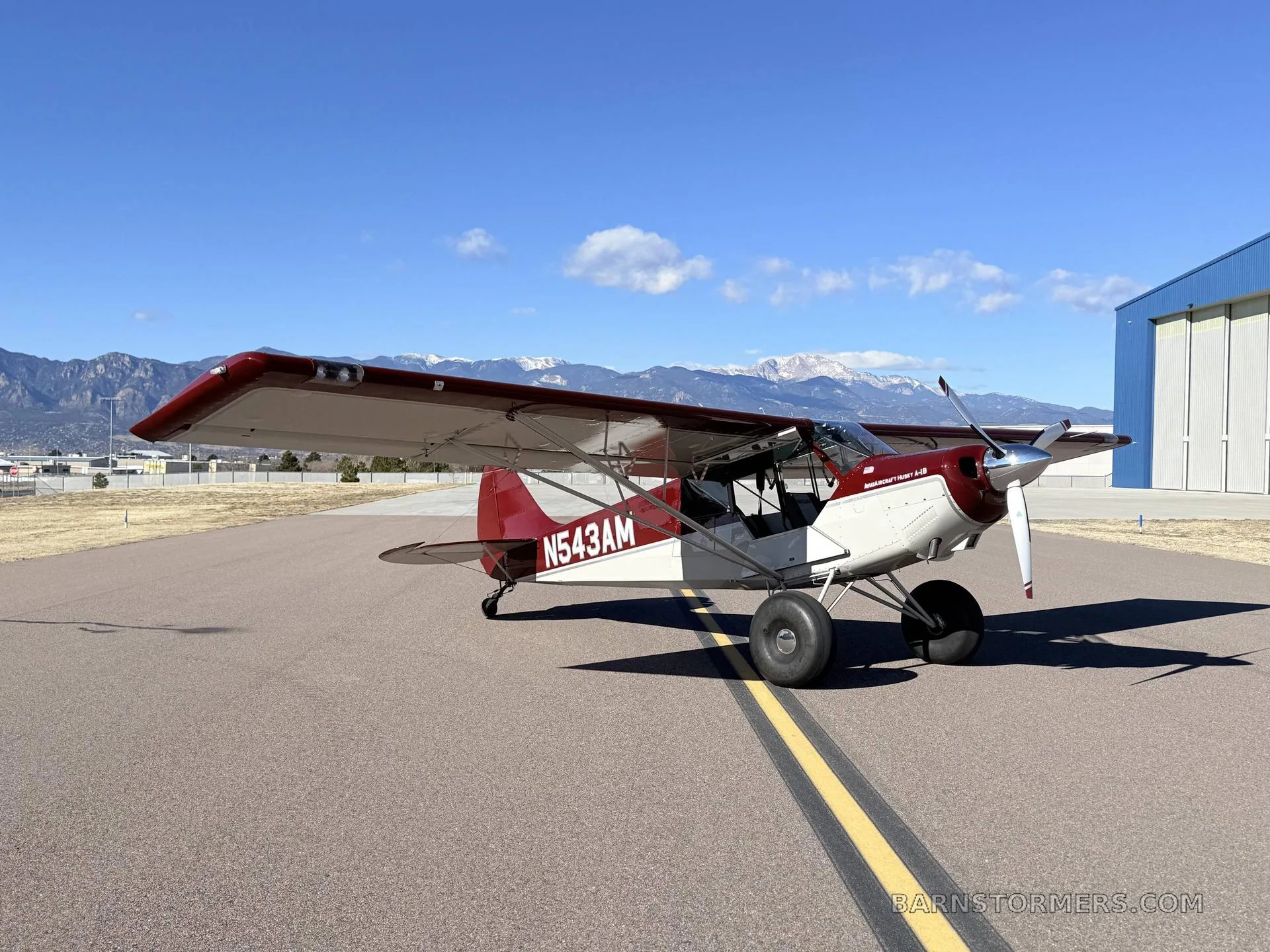 A red and white Aviat Husky A-1B aircraft with large tires is parked on an asphalt runway with mountains in the background.