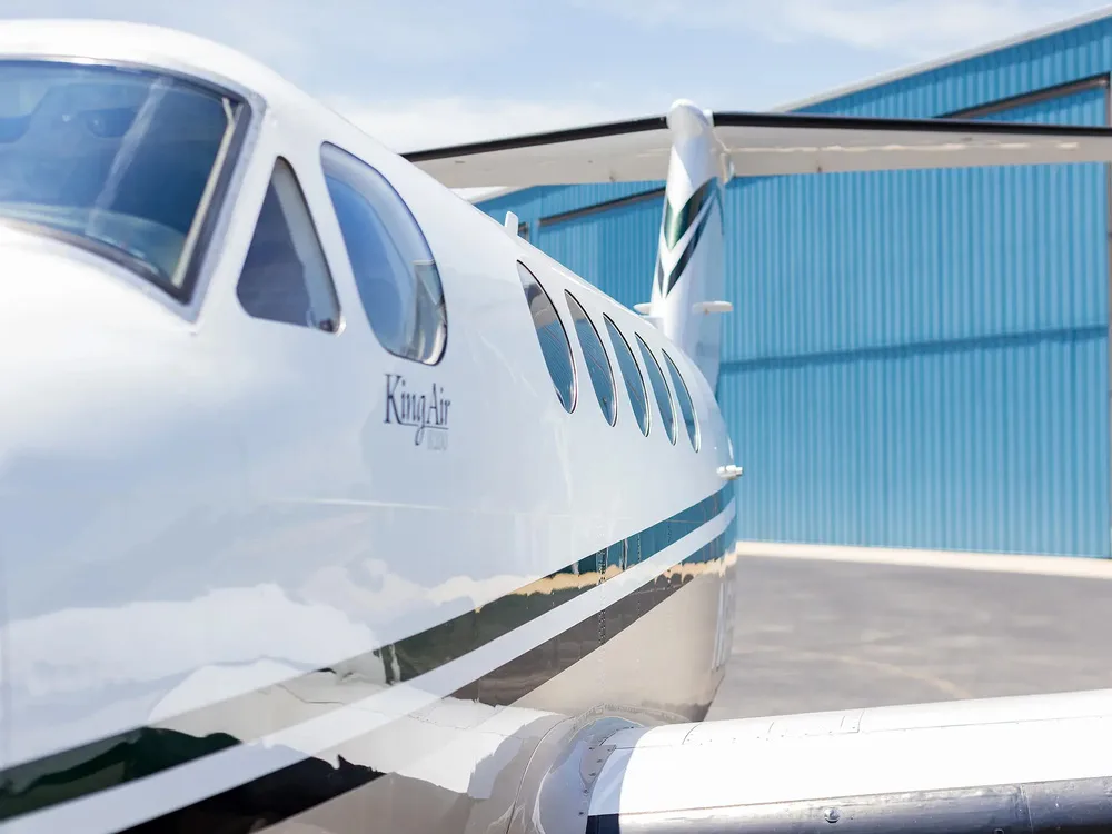 A close-up view of the cockpit windows and forward fuselage of the white Beechcraft King Air, showing its branding and green stripe.