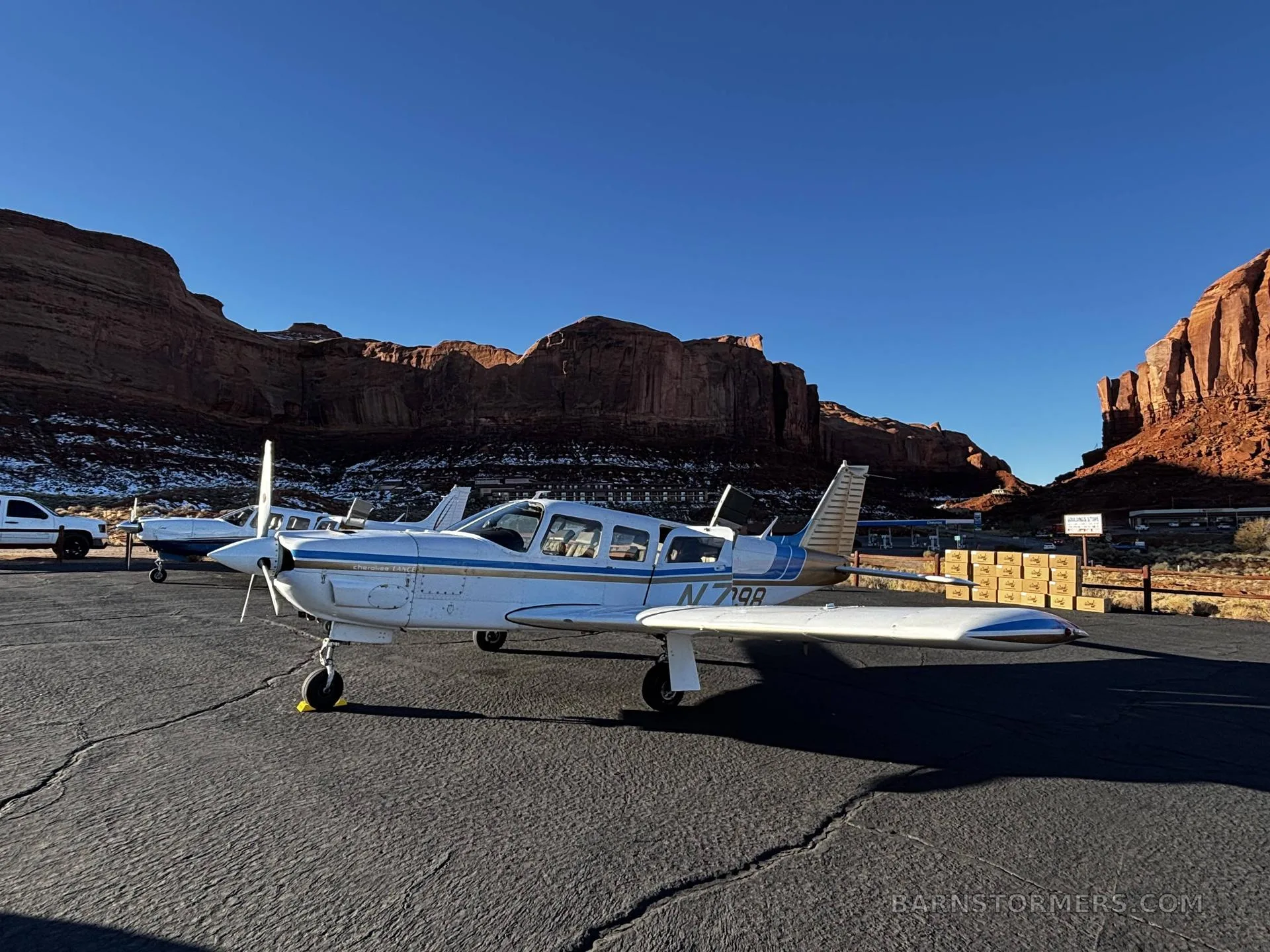 A white and blue Piper Cherokee Lance (N75398) parked on an asphalt ramp with towering red rock formations in the background under a clear sky.
