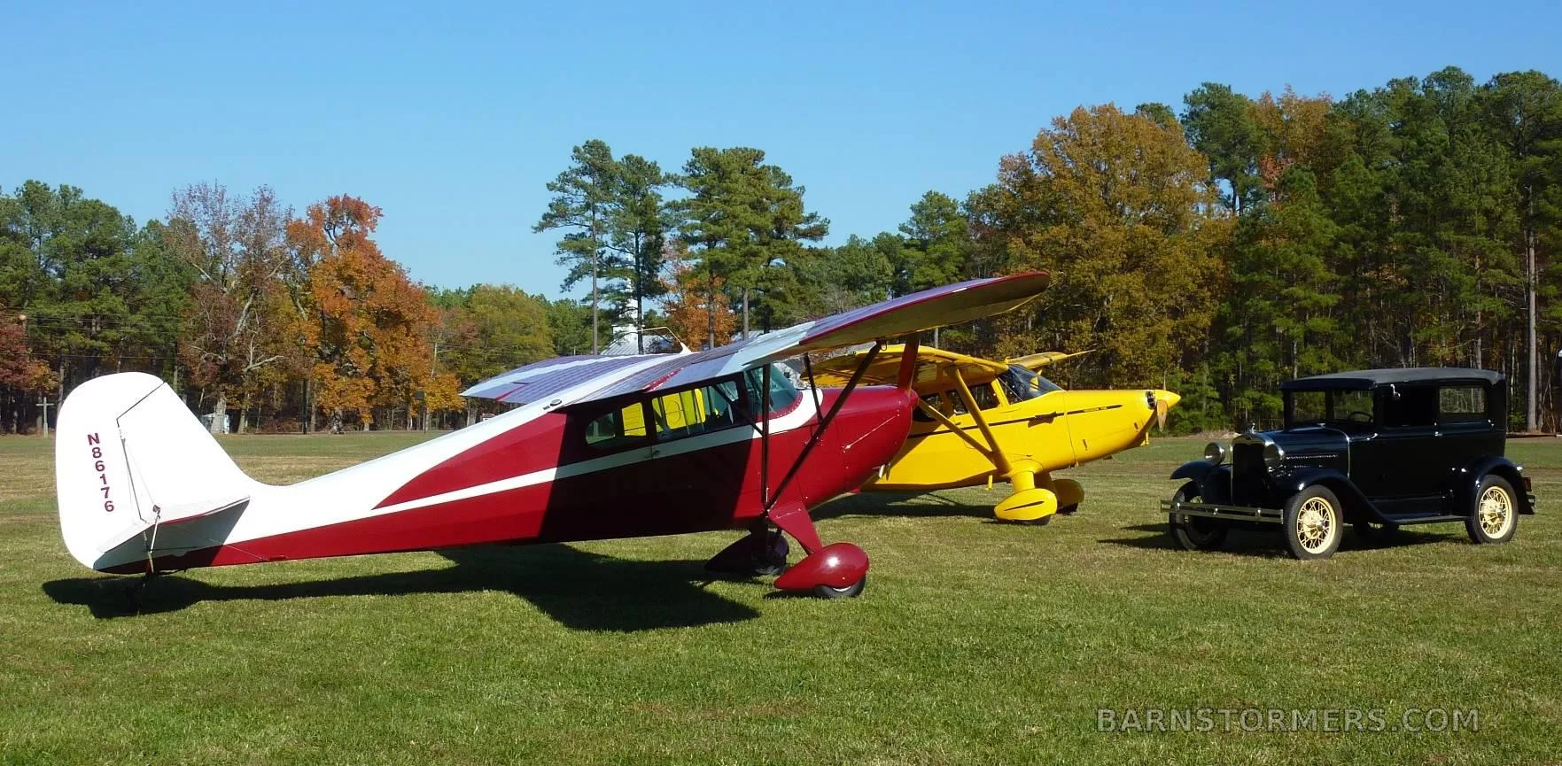 A red and white Aeronca Chief N86176 parked on a grassy field next to a yellow aircraft and a black vintage car, with autumn trees in the background.