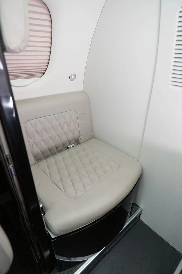 A light gray quilted leather side-facing seat next to a window with closed blinds in the cabin of a business jet.