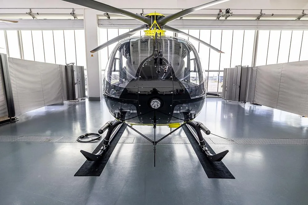 A bright yellow Airbus H135 helicopter is viewed head-on inside a hangar, showing its large front windshield.