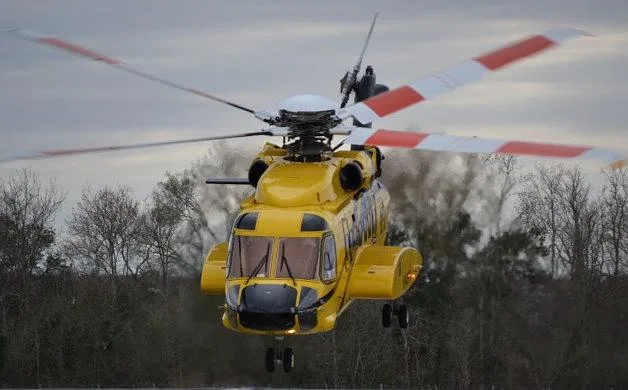 A yellow helicopter with red and white striped rotor blades hovers in flight above a treeline under a cloudy sky.