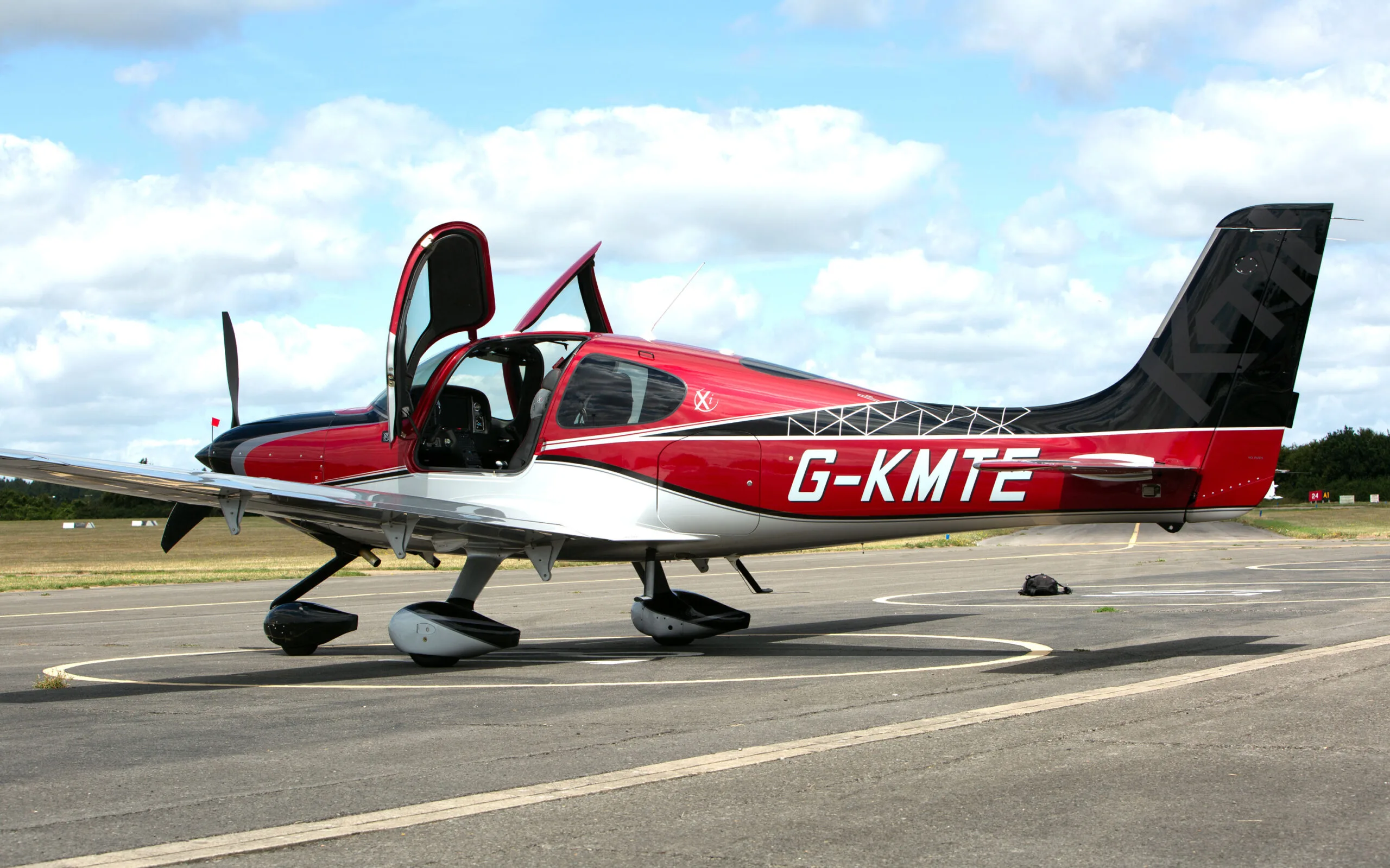 A side view of a red, black, and silver Cirrus SR22 aircraft with registration G-KMTE and open doors, parked on an airfield.