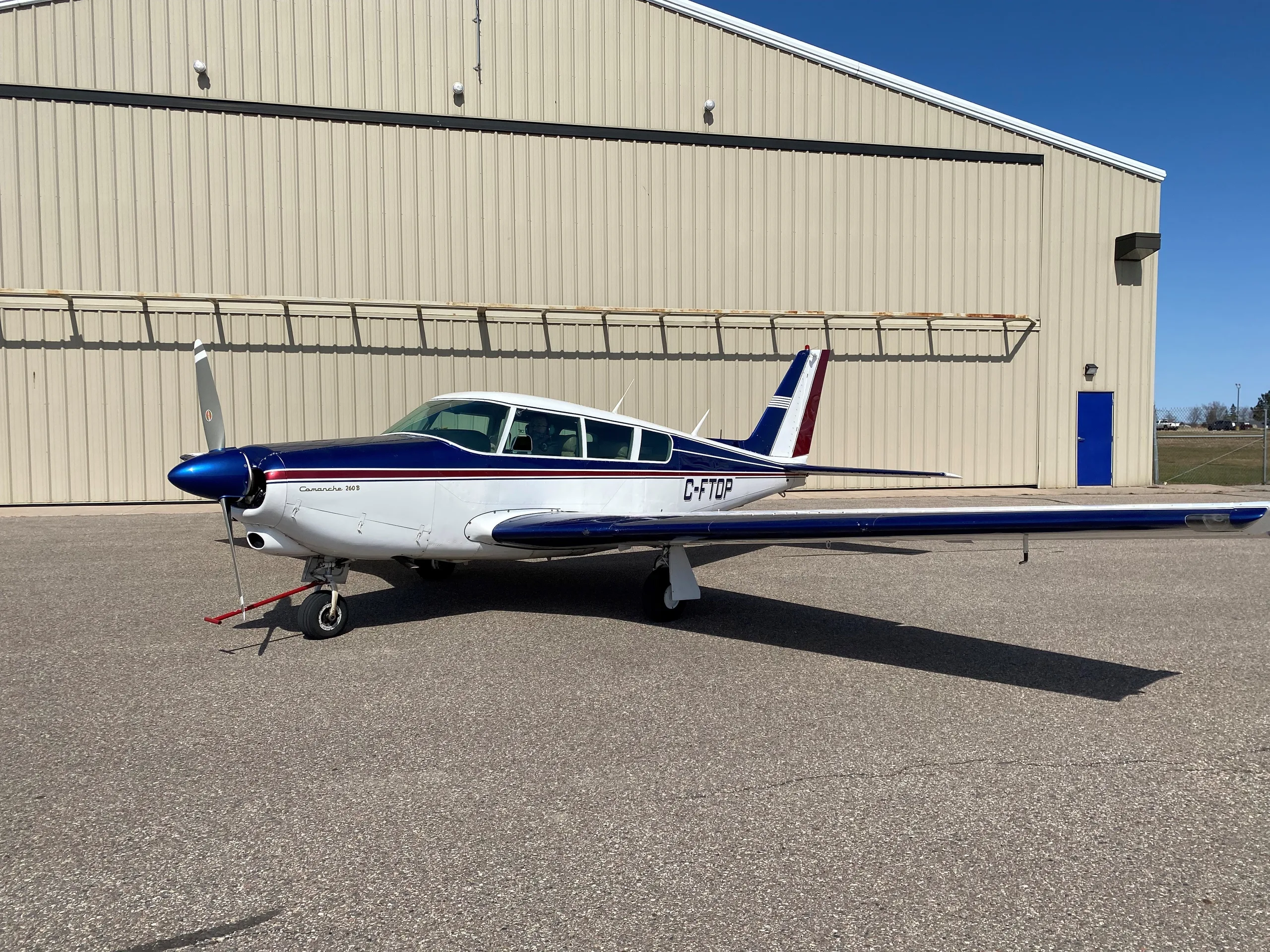 A white Piper Comanche 260B with blue and red stripes is parked on an asphalt ramp in front of a light-colored hangar under a clear blue sky.