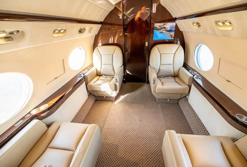 The spacious interior cabin of a private jet with two sets of facing beige leather seats and a screen displaying a map.
