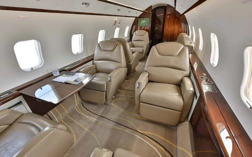 An angled view of the private jet's cabin, featuring tan leather seating, a table, and large windows.
