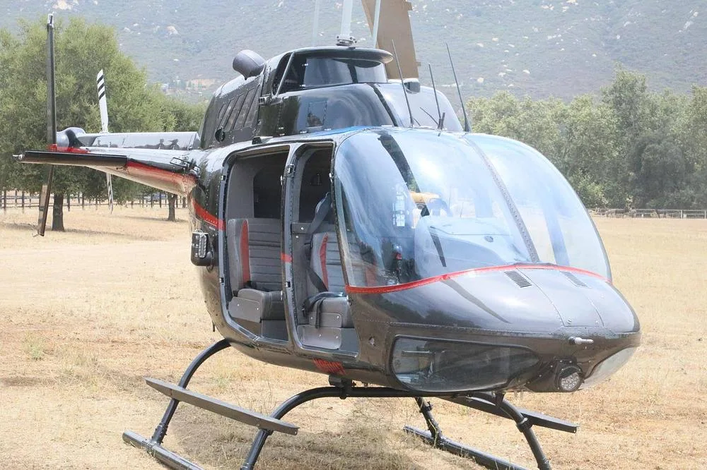 A black helicopter with red trim and open doors is parked on a dry grassy field with trees and hills in the background.