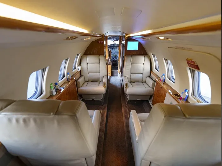 The luxurious interior cabin of a business jet with light tan leather seats and wood paneling, looking towards the cockpit.