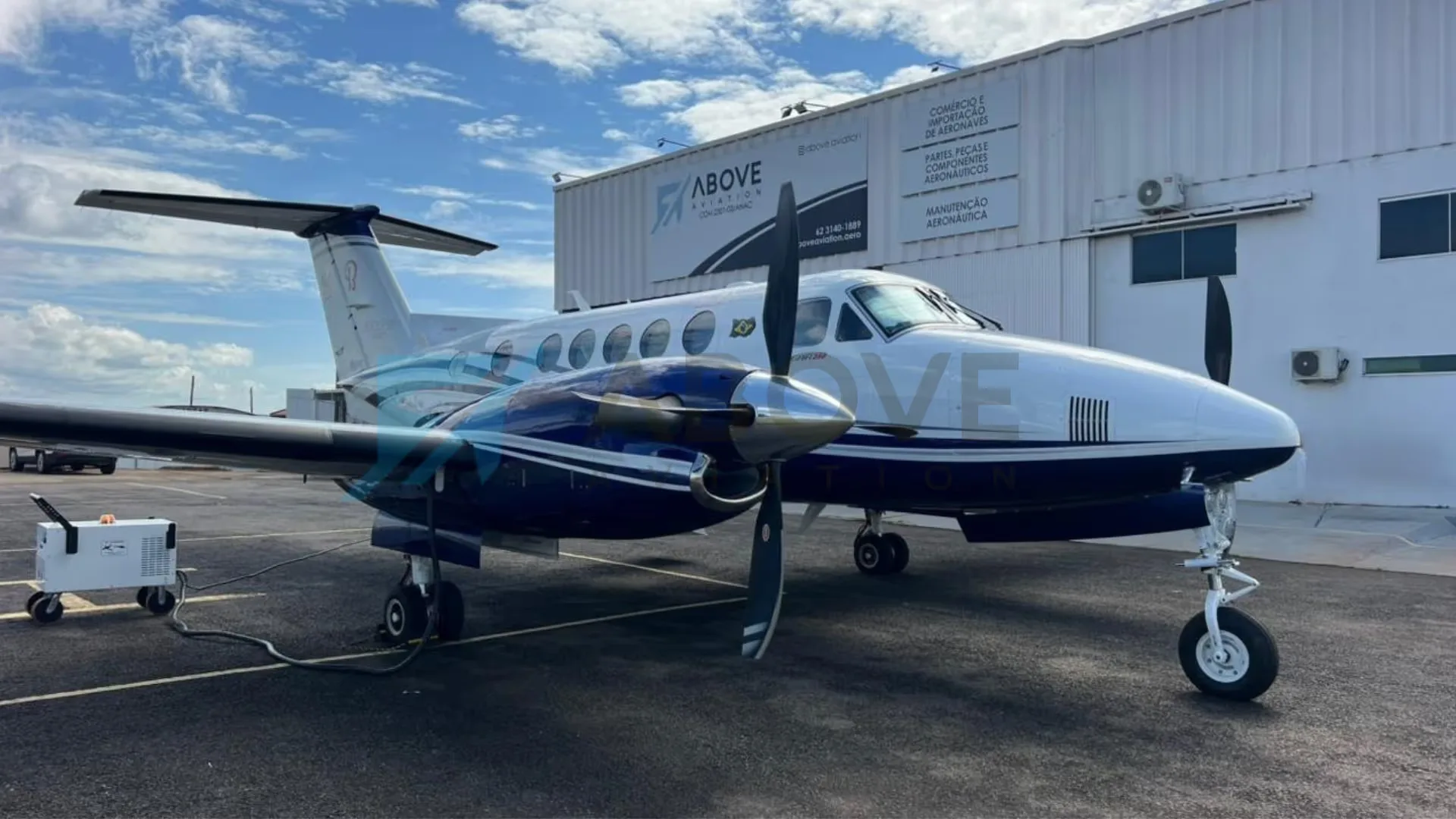 A white and blue Beechcraft King Air turboprop aircraft is parked on an asphalt apron next to a white hangar, with a ground power unit connected.