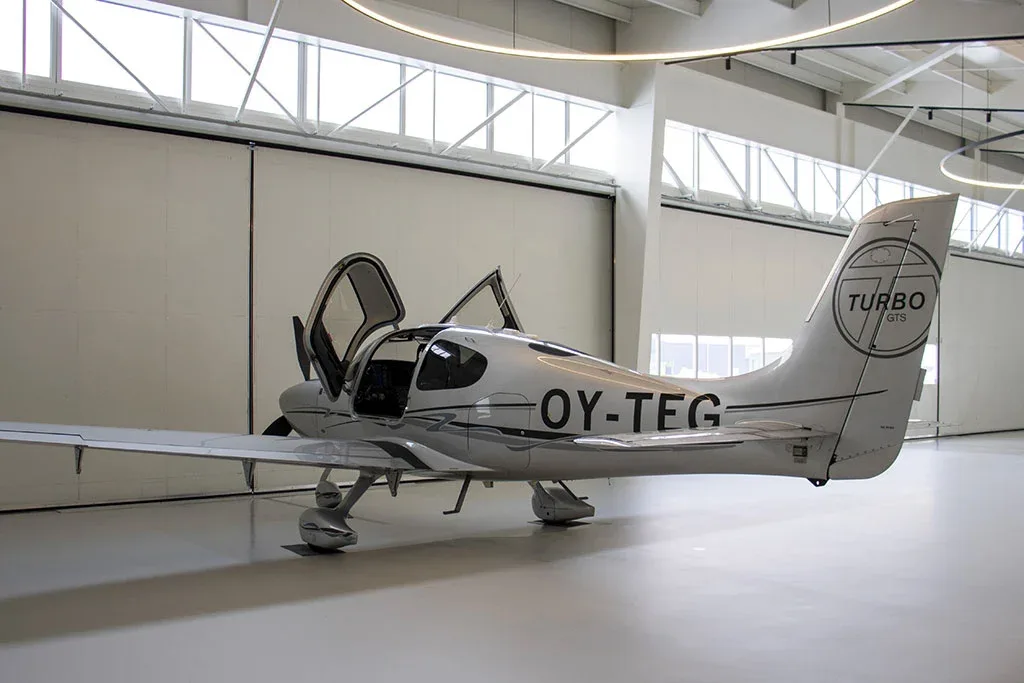 A silver Cirrus SR22 Turbo aircraft, registration OY-TEG, with its doors open, parked inside a modern hangar.