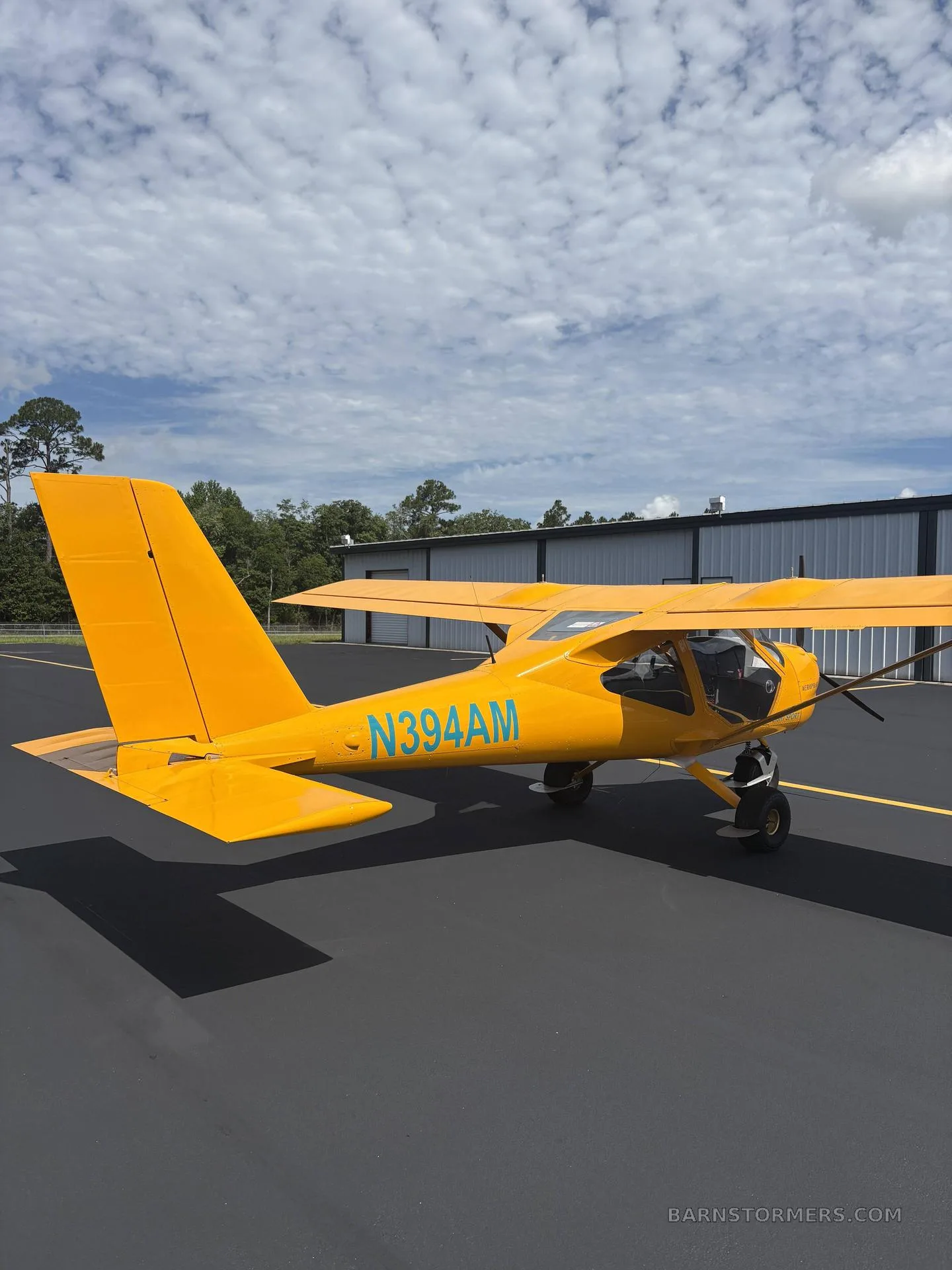 A yellow Aeroprakt A-32 Light Sport aircraft with registration N394AM is parked on an asphalt apron under a partly cloudy sky.