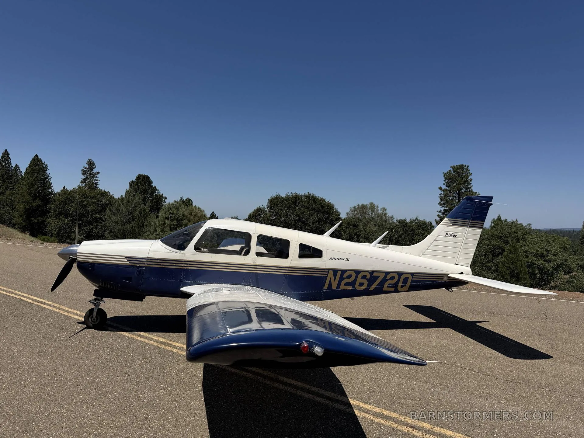 A blue and white Piper Arrow III (N26720) parked on an asphalt surface with trees in the background under a clear blue sky.