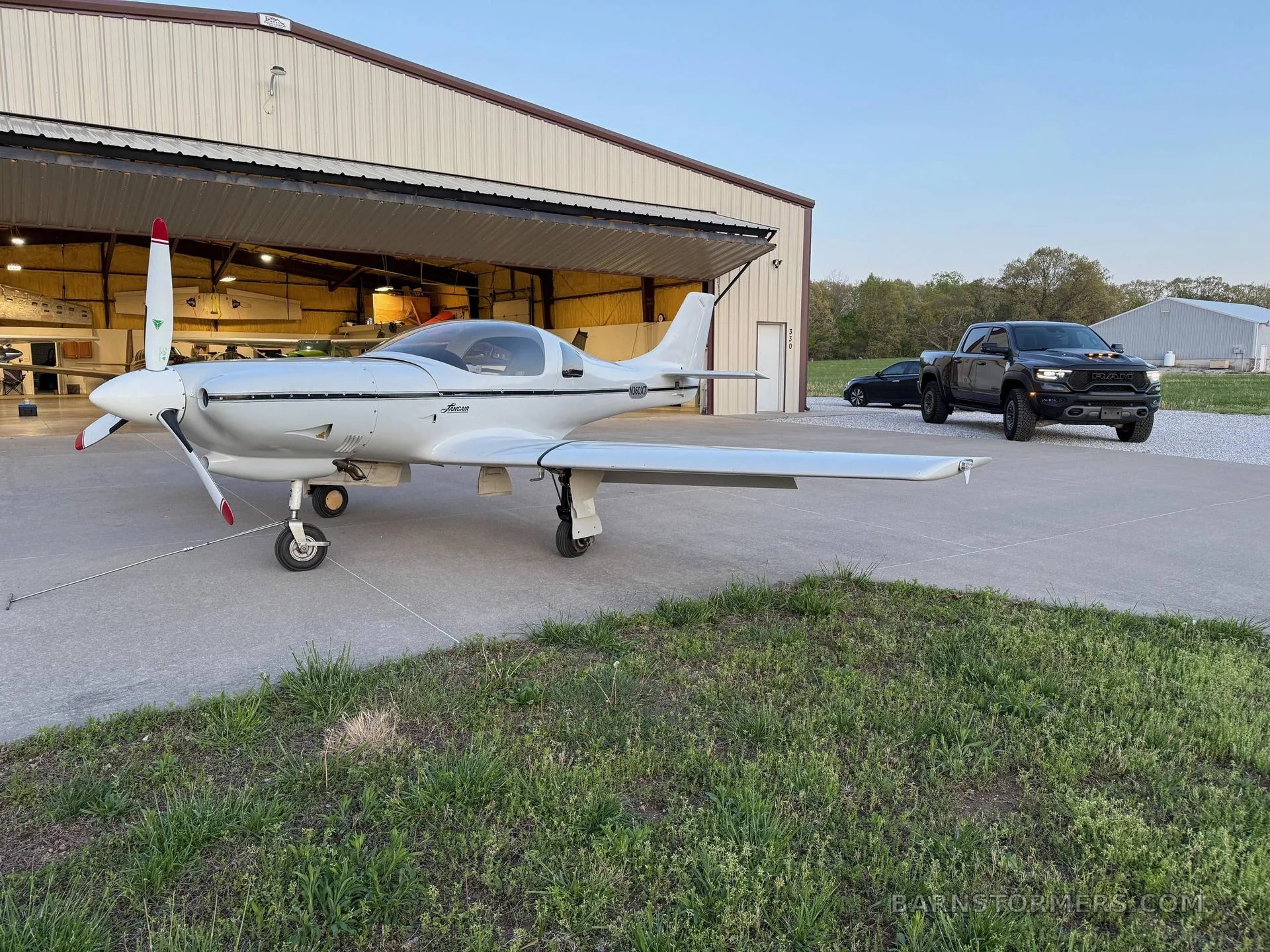 A white Lancair Legacy aircraft with a black stripe is parked on a concrete pad outside a hangar at sunset, with a black pickup truck in the background.