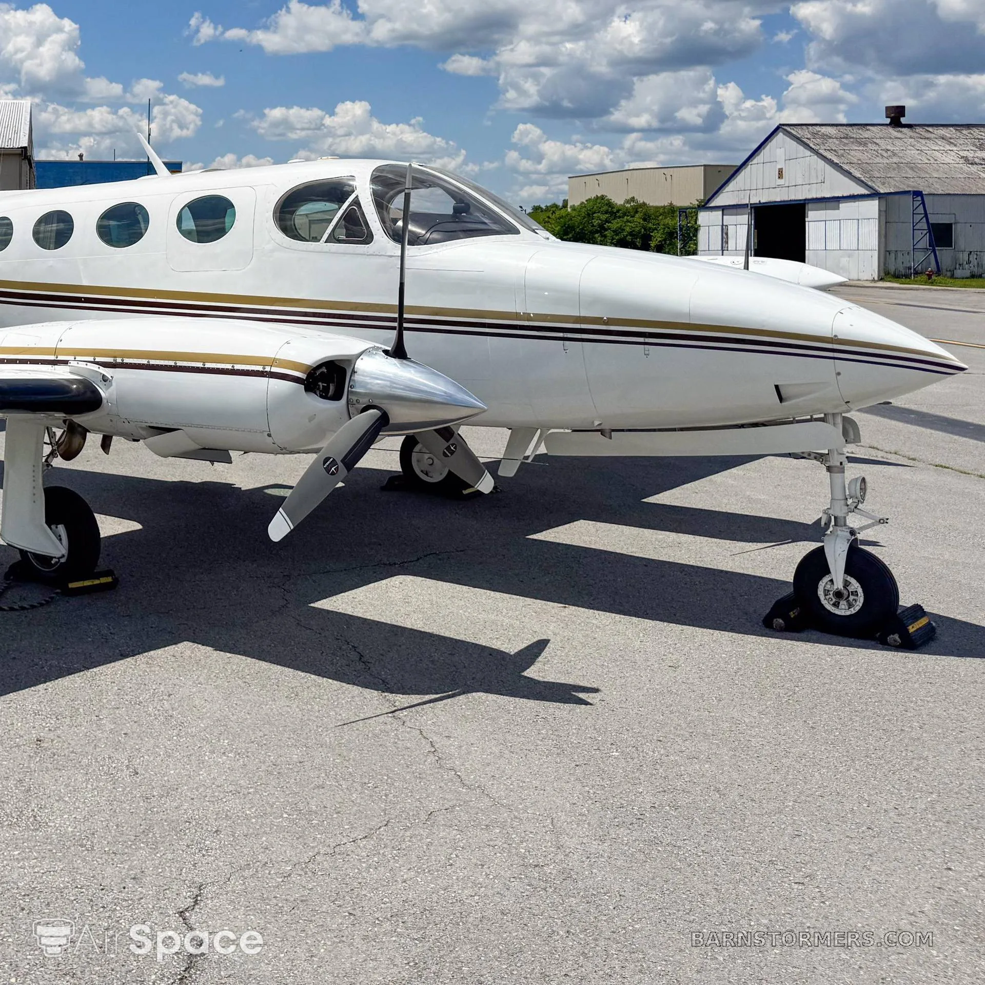 A white twin-engine propeller aircraft with gold and black stripes, parked on an asphalt tarmac with buildings and clouds in the background.